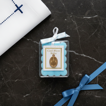 Load image into Gallery viewer, A miraculous medal encased in plastic with a blue bow and cloth and a gold trim. The blessing reads "God Bless Baby". The medal is laying flat on a dark marble surface beside a blue ribbon tied into a bow.