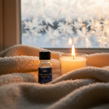 Load image into Gallery viewer, Bottle of 'Cosy Winter Nights' essential oil and a lit candle on a blanket with a frosty window in the background.