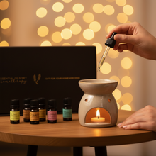 Load image into Gallery viewer, Hand adding essential oil with a dropper to a ceramic burner beside a lit candle, with the Refresh &amp; Revive aromatherapy gift set in the background and warm bokeh lights for a calming night routine.
